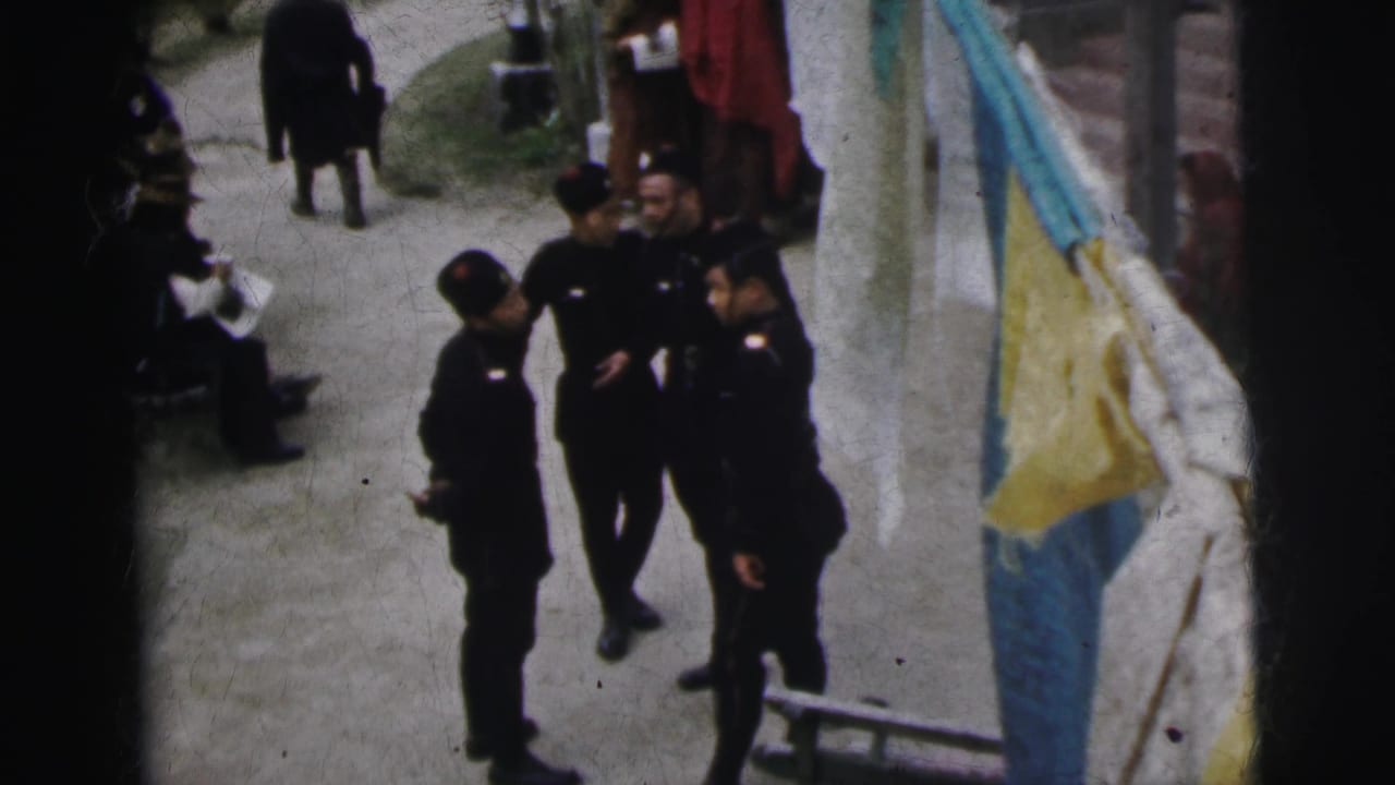 8mm home movie frame, India 1962: several people in dark coats near hanging yellow and teal fabric on a clothesline, outdoor dirt-path setting with trees behind.