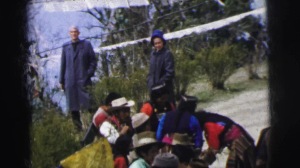 8mm film frame, India 1962: an elderly bald man and a woman in long blue overcoats observe a group wearing wide-brimmed hats and layered colorful garments on a hillside.