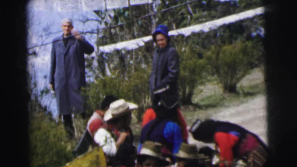 8mm home movie, India 1962: older man in blue-gray coat and woman in blue headscarf stand above a group wearing wide-brimmed hats and multilayered colorful garments on a hillside path.