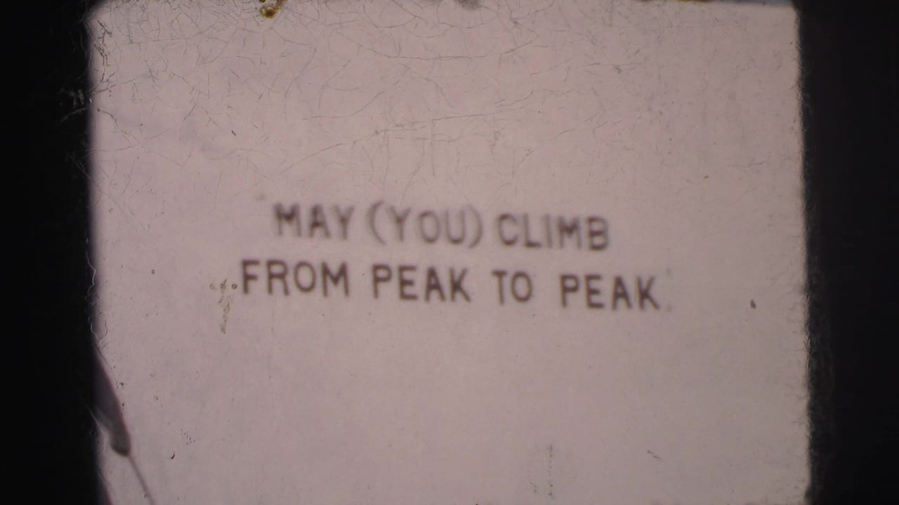 1962 Calcutta home-movie frame showing a faded peak-themed sign with the phrase MAY (YOU) CLIMB FROM PEAK TO PEAK.