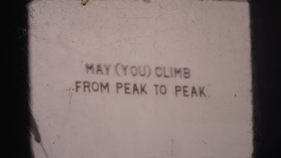 1962 Calcutta home-movie frame showing a faded peak-themed sign with the phrase MAY (YOU) CLIMB FROM PEAK TO PEAK.