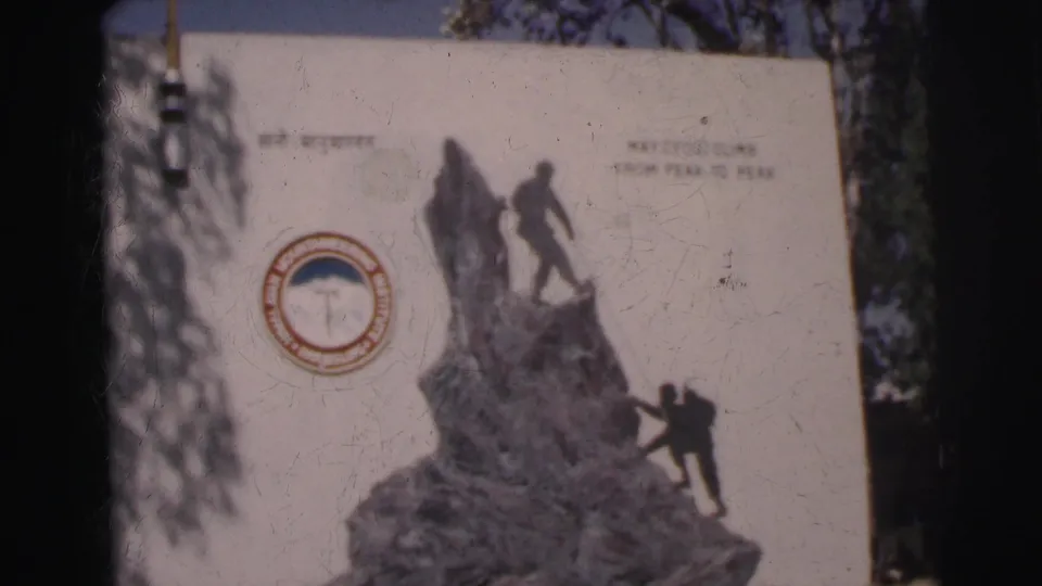 1962 India home-movie frame showing a climbing monument panel with HMI logo, climber figures, and the motto about climbing peak to peak.