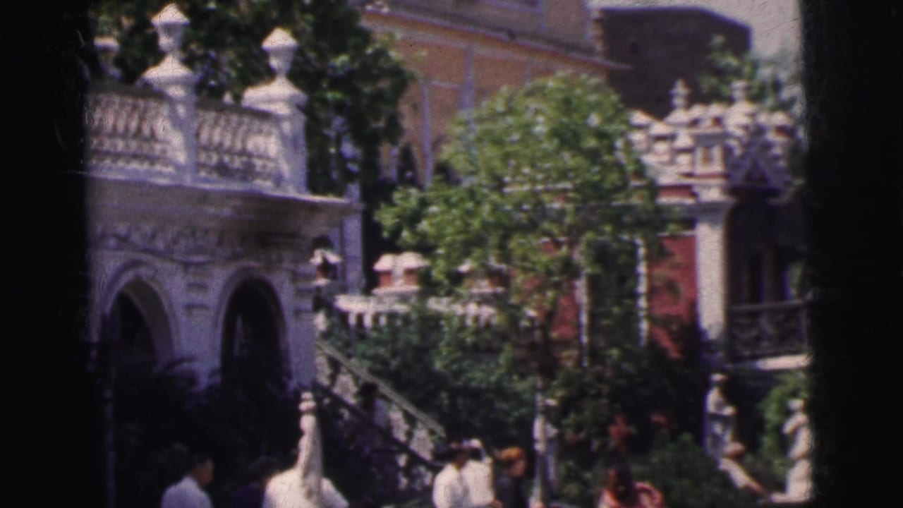 Ornate white balustrade staircase and arched building facade with adult pedestrians passing, 8mm film.