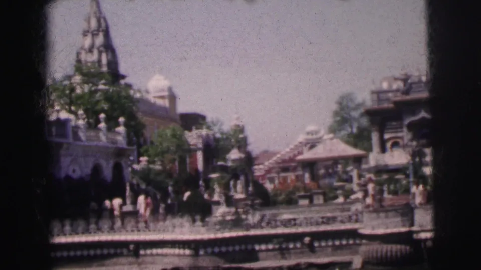 Wide 8mm shot of a Calcutta street precinct showing temple spires, ornate pavilion structures, decorative railings, and adult pedestrians against a pale sky, circa 1962.