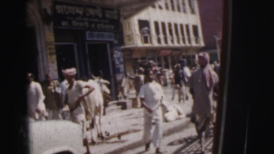 Calcutta, India, c. 1962 — men in white traditional clothing and a cow share the pavement outside a Bengali-script shop; colonial facade behind.