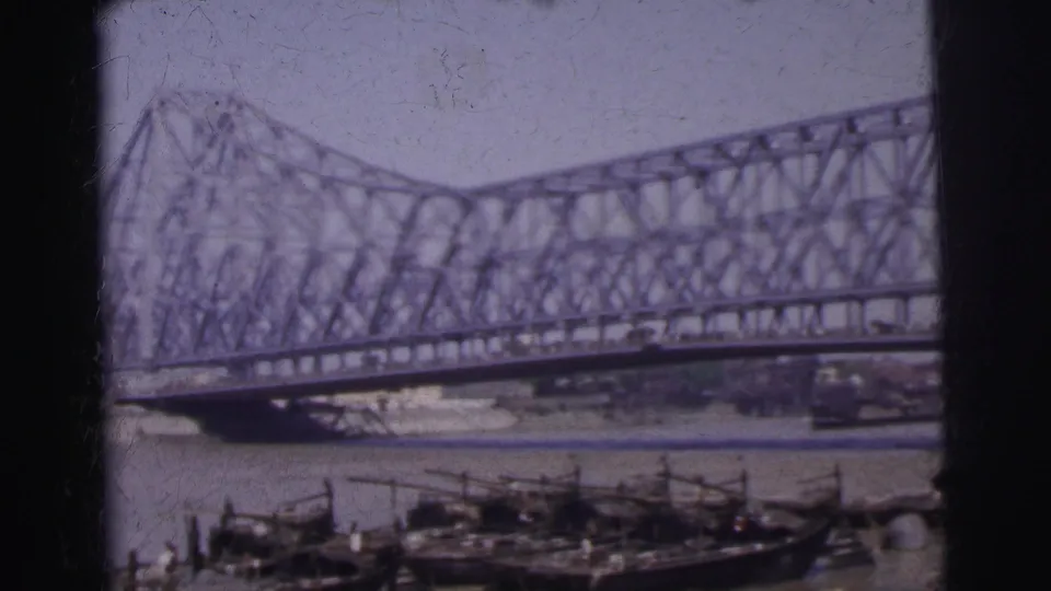 A steel bridge spans water with small boats below, appearing as urban context within the Calcutta 1962 reel.