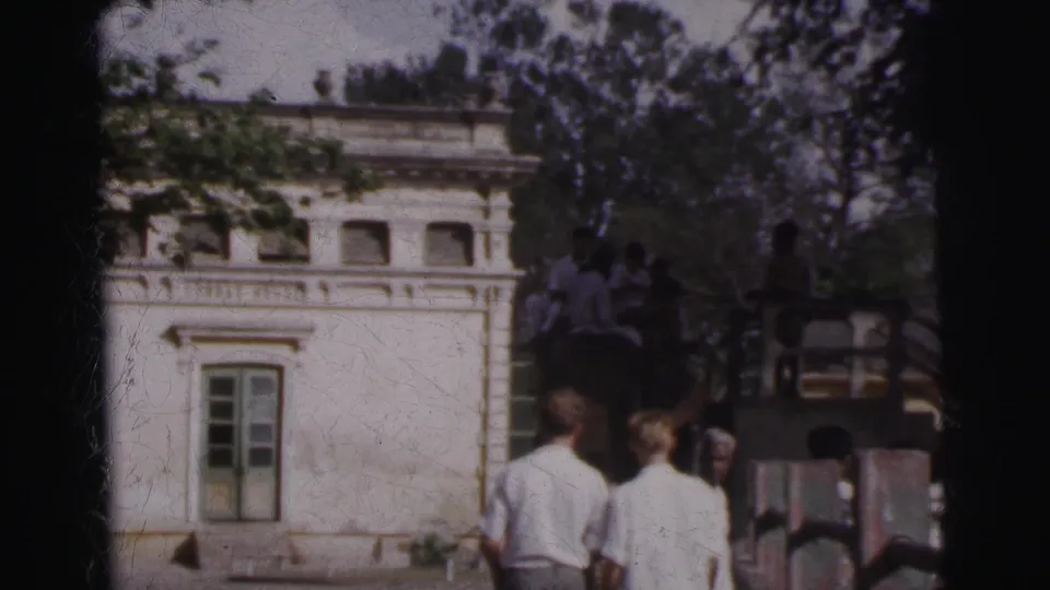1962 Calcutta home-movie frame showing a white colonial-style building, trees, visitors, and a vehicle near the scene.