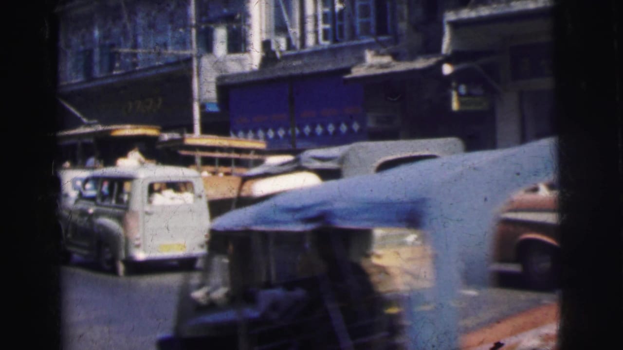 Busy street with a trolley, white van, rickshaw-like vehicle, and storefronts.