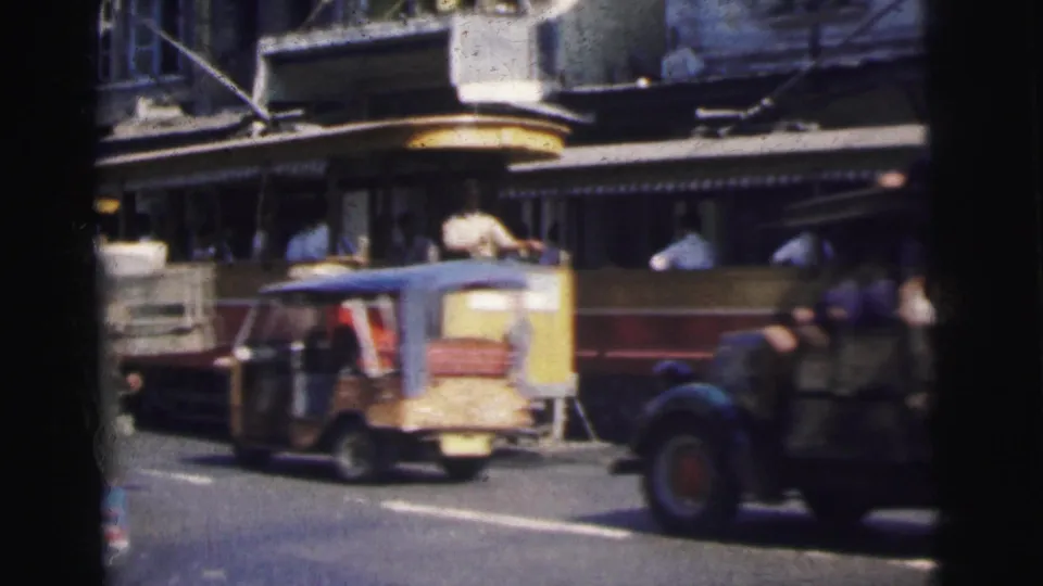 A 1962 Calcutta trolley moves through mixed traffic, with passengers visible and a dark vehicle crossing the foreground.