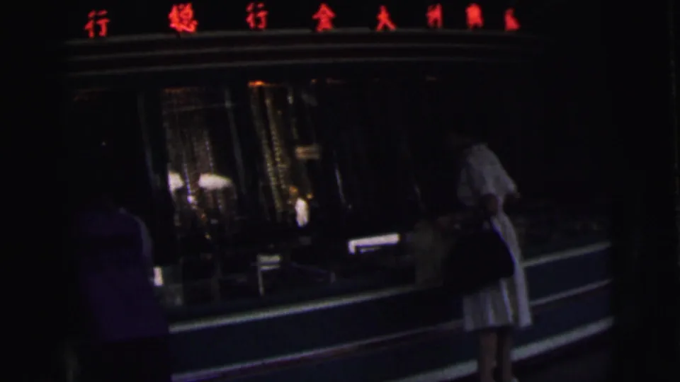 Dark 8mm frame of a Calcutta storefront in 1962, with a woman in white and another figure near a glass display beneath red Chinese characters.