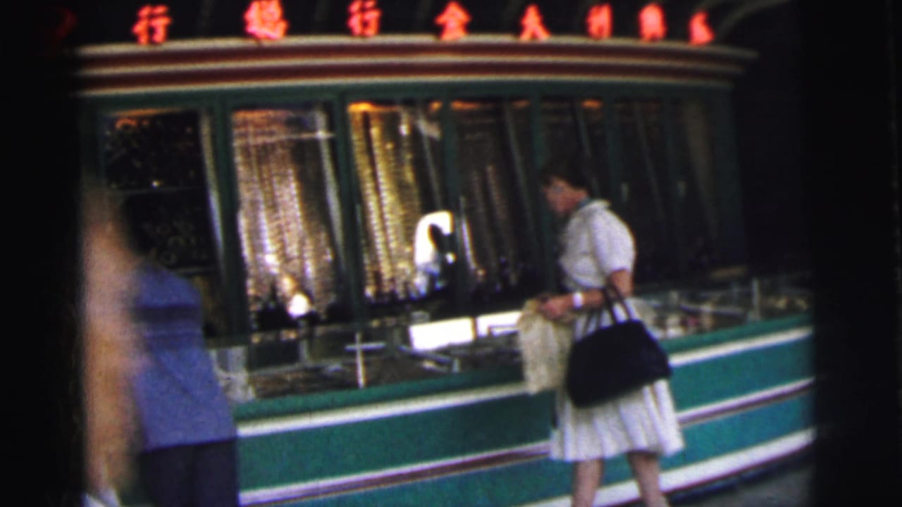 In 1962 Calcutta, a woman in a white dress carries a black handbag while looking at a green-and-white storefront with red Chinese characters above.