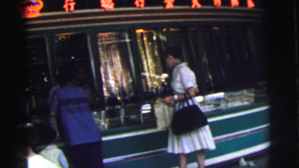 A man in a blue shirt and a woman in a white dress stand at a Calcutta shopfront in 1962, with red Chinese characters above the window.