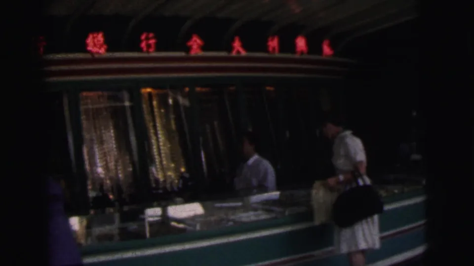 1962 Calcutta home-movie frame showing a dark shop or counter area with red neon signage and shoppers partly visible.