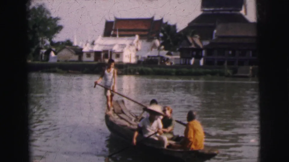 Home-movie frame from Calcutta in 1962 showing a wooden paddle boat, passengers, and soft-focused buildings along the opposite bank.