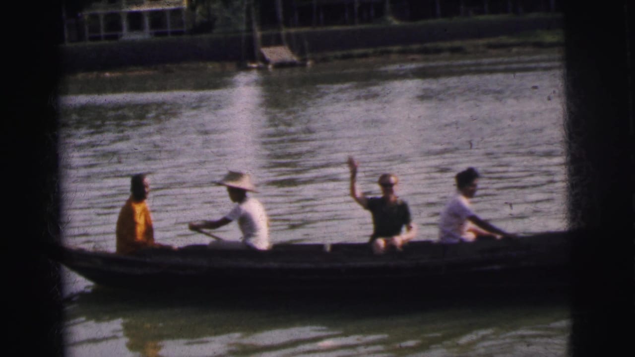 1962 Calcutta home-movie frame showing four adults in a long canoe, a paddler in a broad hat, and one passenger waving on the river.