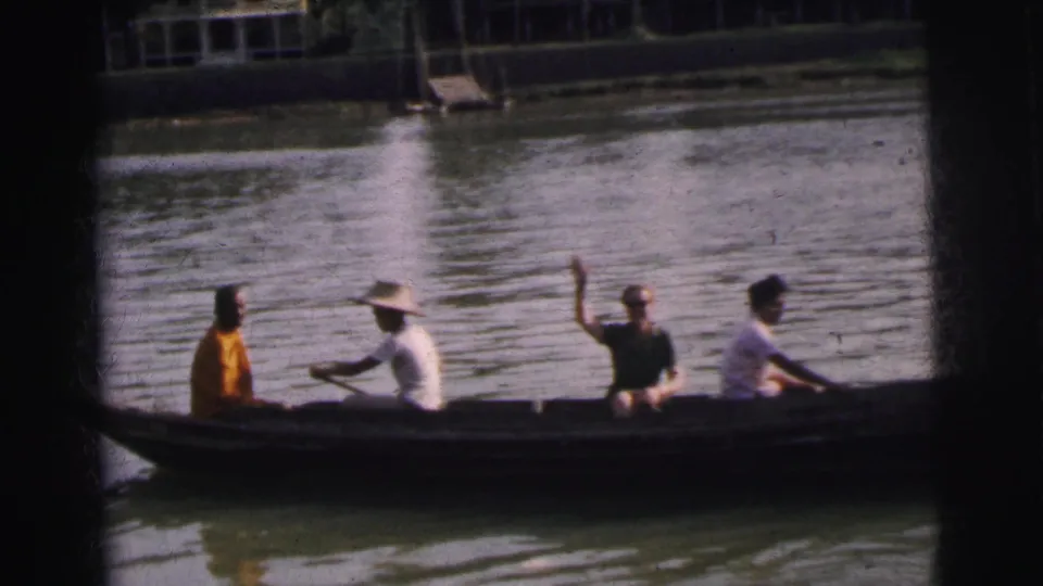 1962 Calcutta home-movie frame showing four adults in a long canoe, a paddler in a broad hat, and one passenger waving on the river.