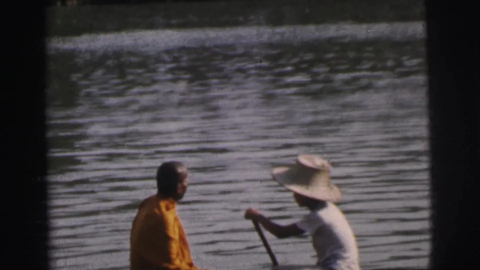 Close 8mm frame from Calcutta showing an orange-robed passenger and a broad-hatted paddler seated in a long canoe on calm water.
