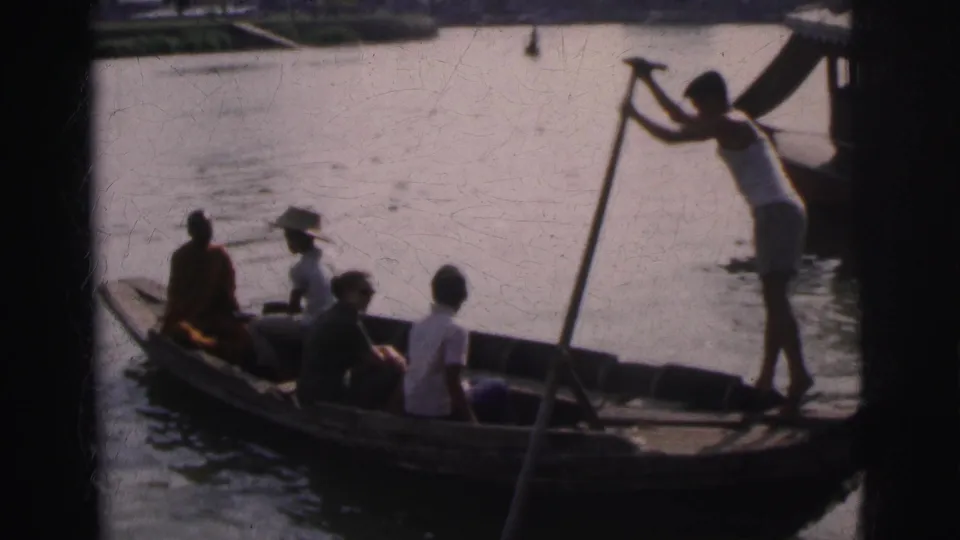 Boatman stands and paddles a wooden passenger boat in 1962 Calcutta, with seated travelers and a robed figure aboard.
