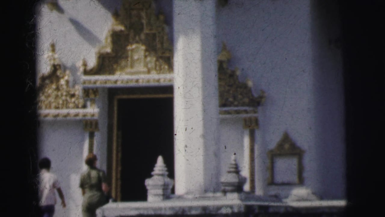 Two visitors approach a white temple entrance with ornate gilded decoration and columns.