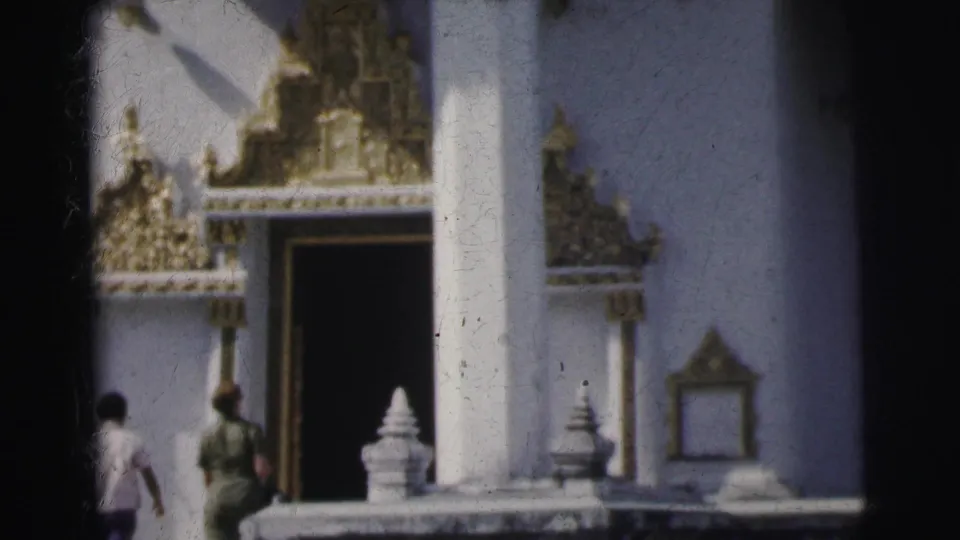 1962 Calcutta home-movie frame showing two visitors walking toward a dark doorway in a white temple facade with gilded ornament.