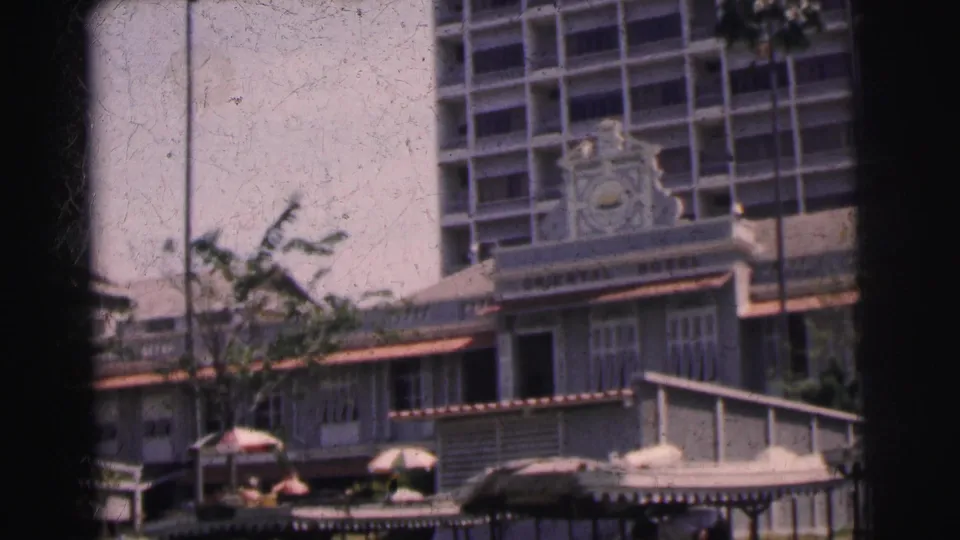1962 Calcutta 8mm frame showing the Oriental Hotel facade above a street frontage with umbrellas, trees, awnings, and distant people.