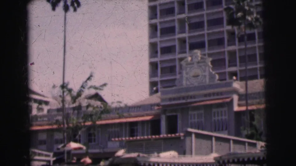 1962 Calcutta 8mm frame showing the Oriental Hotel building from the street, with awnings, umbrellas, trees, and small figures below.