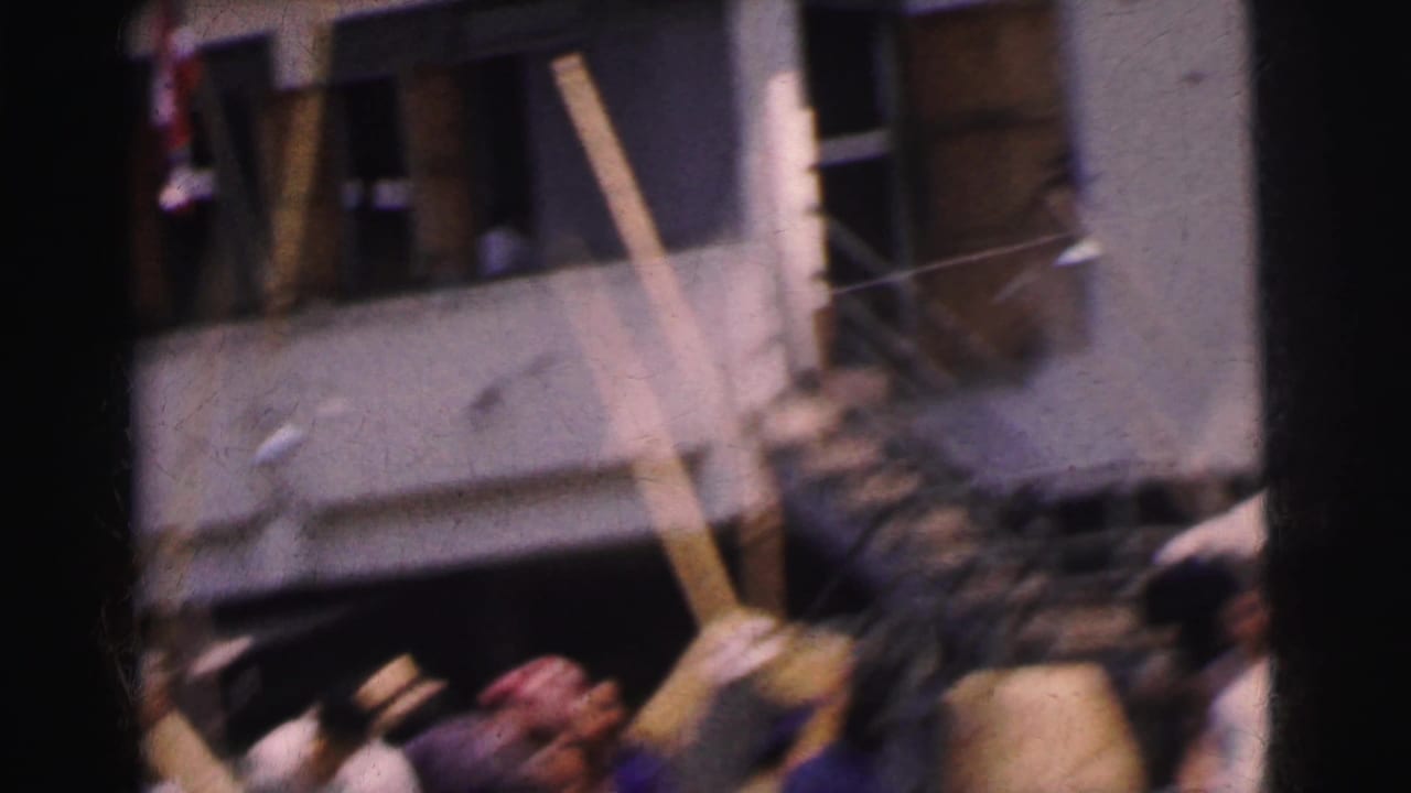 Handheld 1962 Calcutta street view with a blurred crowd, umbrellas or bundles, and a building stairway in the background.