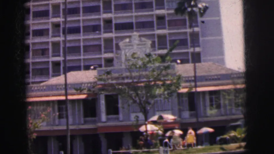 1962 Calcutta frame showing a colonial-style street facade, trees, umbrellas, and pedestrians beneath a taller modern building.