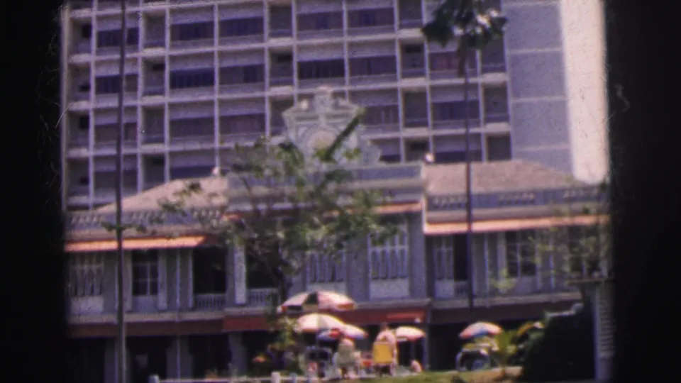 1962 Calcutta home-movie frame with pedestrians under umbrellas, street trees, a colonial-style facade, and a taller building behind.