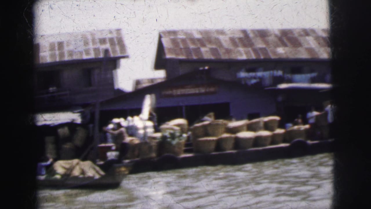Boats loaded with baskets beside wooden riverside buildings and corrugated roofs.