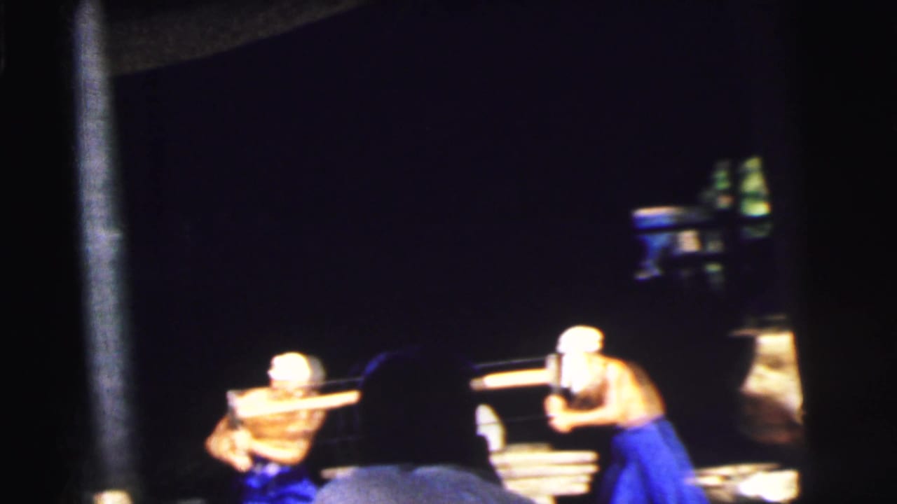 In 1962 Calcutta footage, men in white hats appear to cut or handle wood on a boat viewed through dark foreground framing.