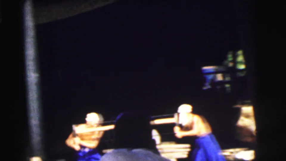 In 1962 Calcutta footage, men in white hats appear to cut or handle wood on a boat viewed through dark foreground framing.