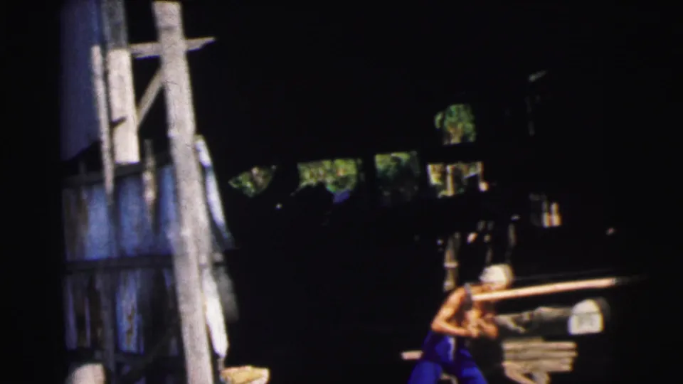 1962 Calcutta home-movie frame showing two men working in a shadowed shed, pulling a long saw across timber.