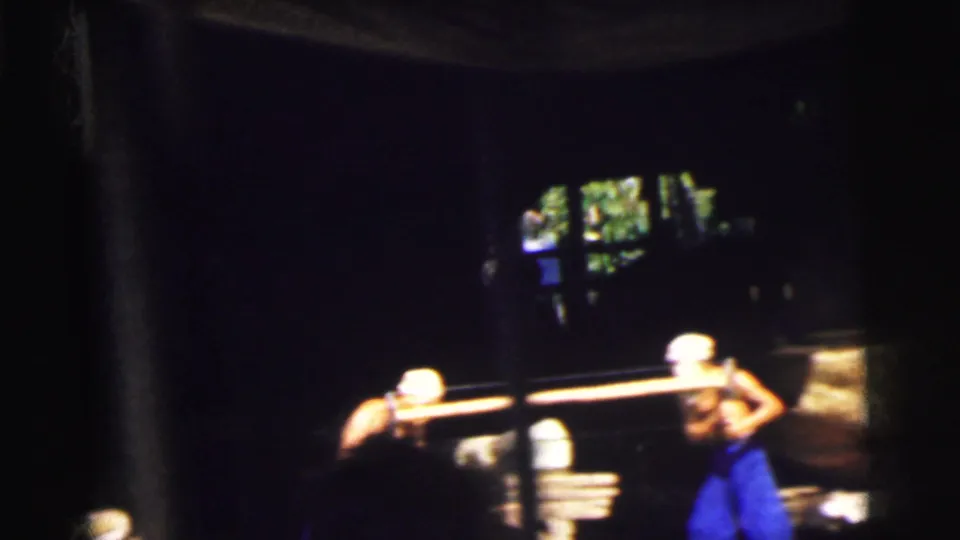 1962 Calcutta 8mm frame of two men in blue trousers sawing timber inside a dim work shed near an open side.
