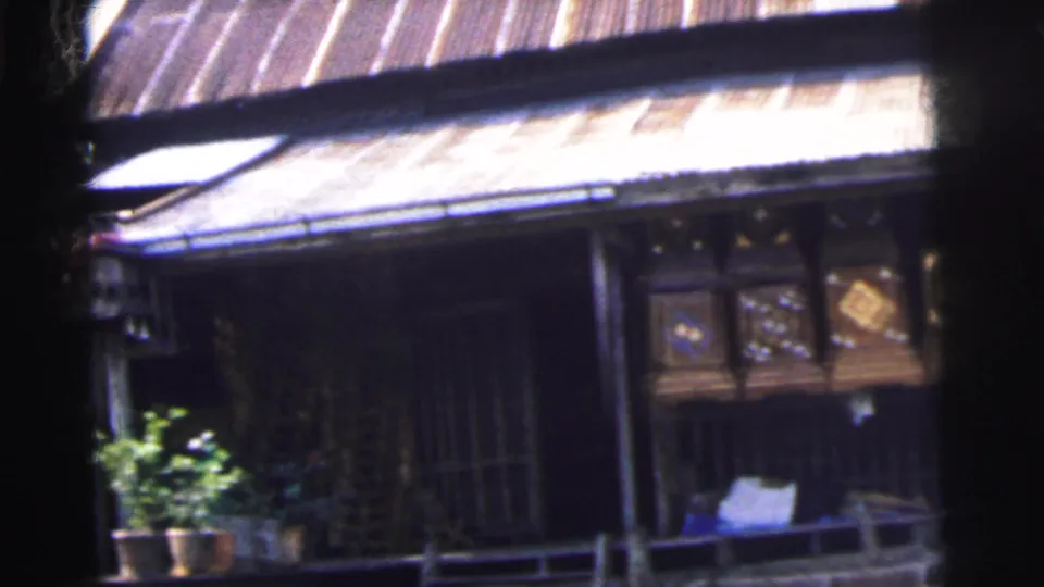Handheld 1962 Calcutta frame showing the boathouse facade at an angle, with decorative panels, plants, and shaded openings.