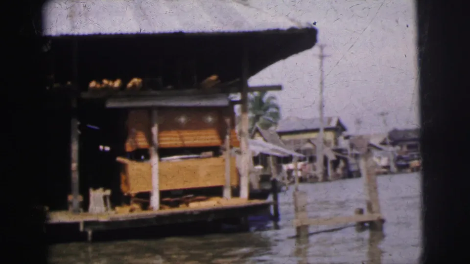 1962 Calcutta frame showing a wooden riverside shop on stilts, water in front, and small structures beyond.