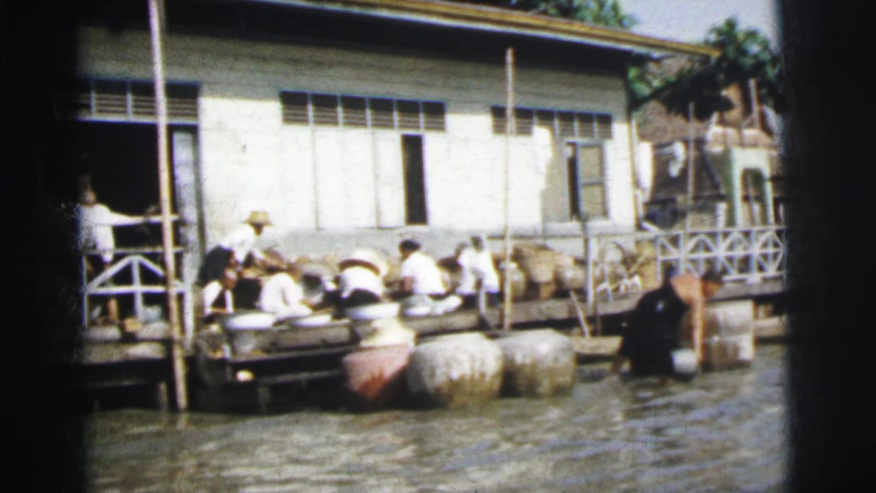 In 1962 Calcutta, workers sit along a wooden riverside dock near large jars, water, and a white building with louvered windows.