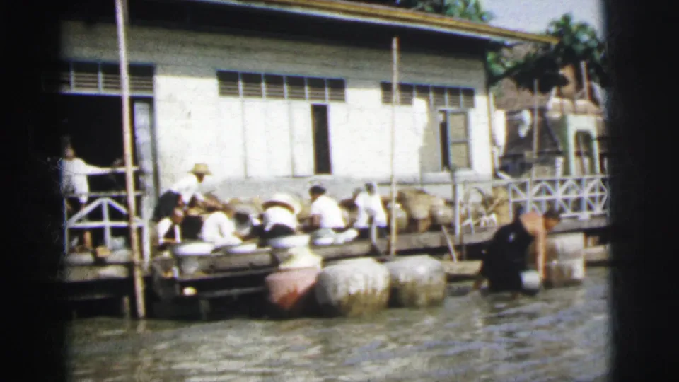 In 1962 Calcutta, workers sit along a wooden riverside dock near large jars, water, and a white building with louvered windows.