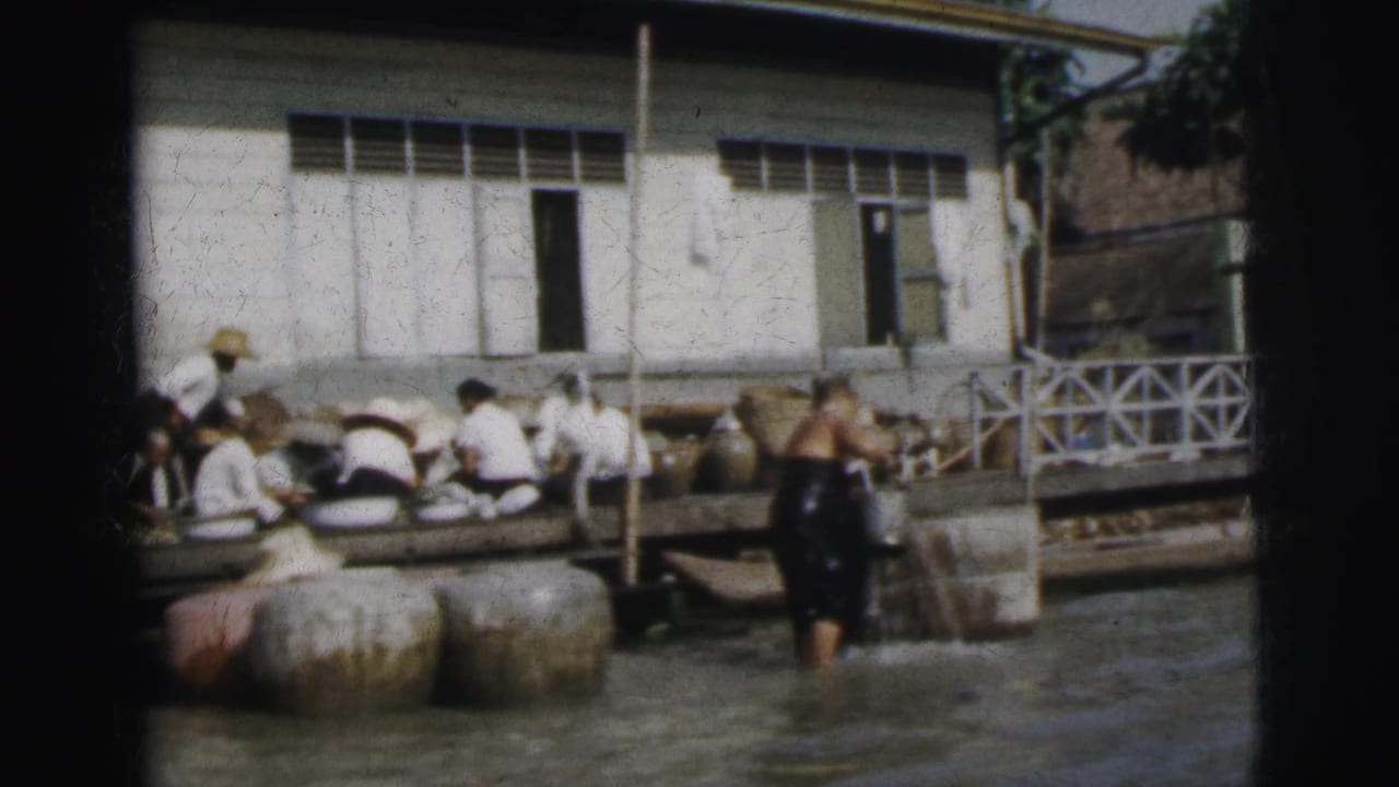 In 1962 Calcutta, a man in dark clothing stands in shallow water near seated dock workers, large jars, and a white riverside building.