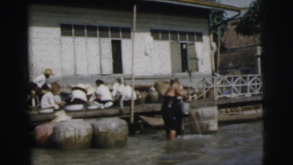 In 1962 Calcutta, a man in dark clothing stands in shallow water near seated dock workers, large jars, and a white riverside building.