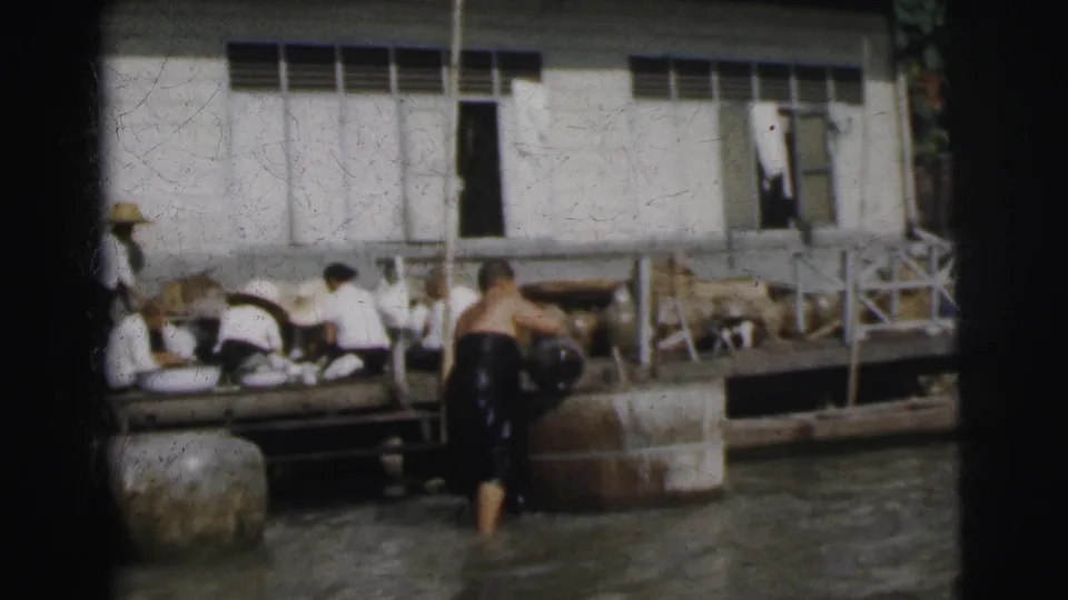 In 1962 Calcutta, seated workers remain on the dock as a man stands in the water near large jars and a white building.