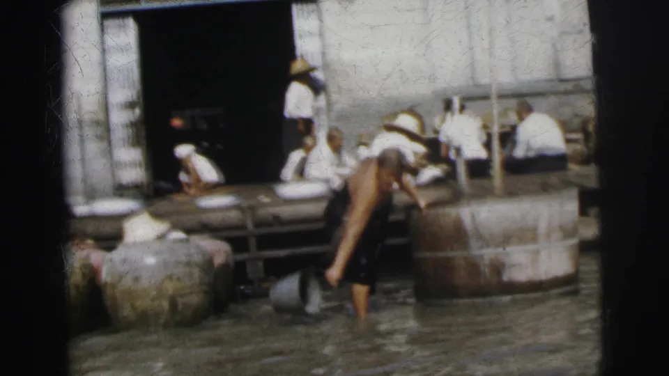 In 1962 Calcutta, a man bends near large jars in shallow water while workers sit on a dock outside an open doorway.
