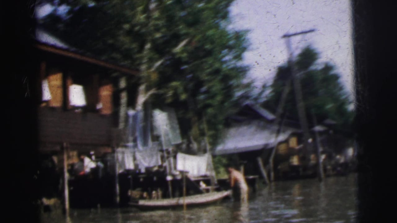 1962 Calcutta home-movie frame showing raised wooden houses, laundry, floodwater, and a small boat along the waterway.