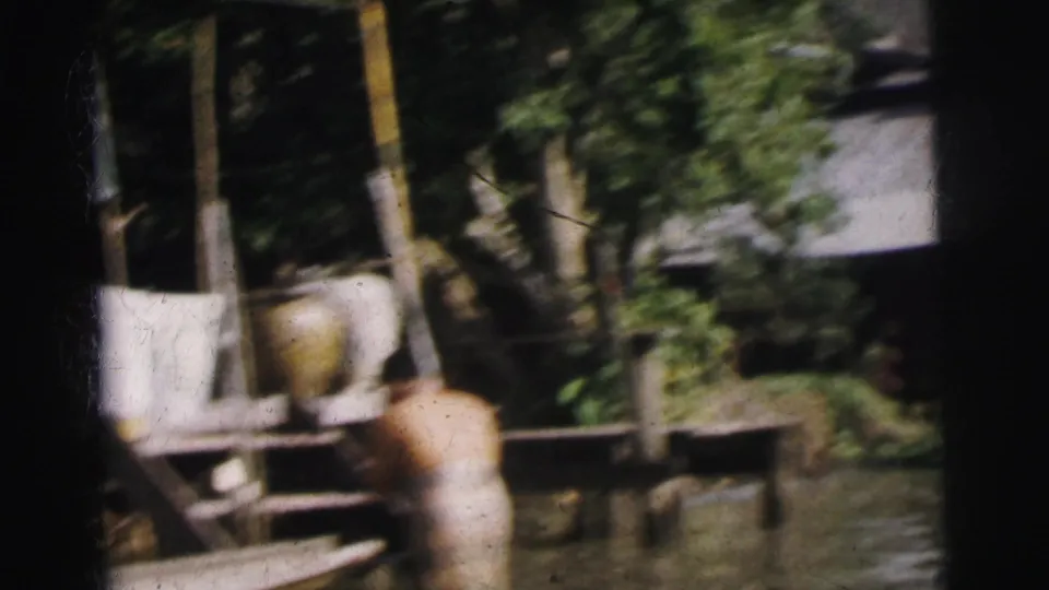 1962 Calcutta home-movie view of an adult by floodwater below raised structures, trees, and hanging laundry.