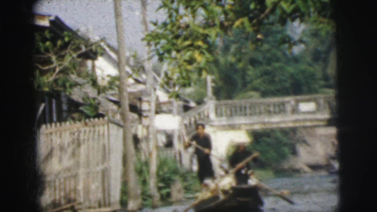 Two people in a small boat move along a narrow waterway beside fences and trees.