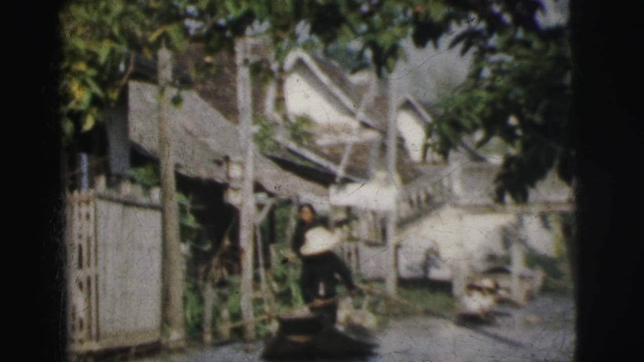 1962 Calcutta frame with a person standing in a narrow boat near a fenced riverside settlement and leafy trees.