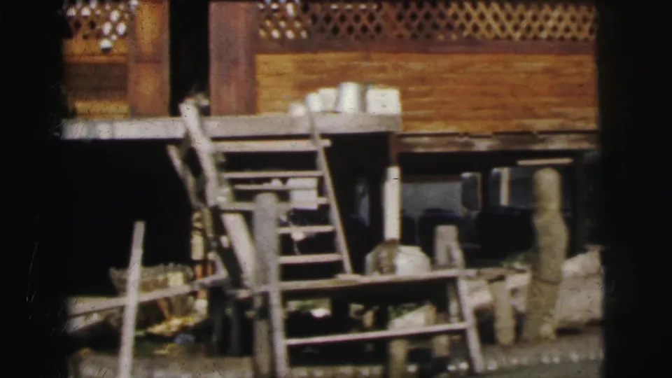1962 Calcutta home-movie frame showing a wooden stilt-like waterside structure, steps, posts, and stored objects near the channel.