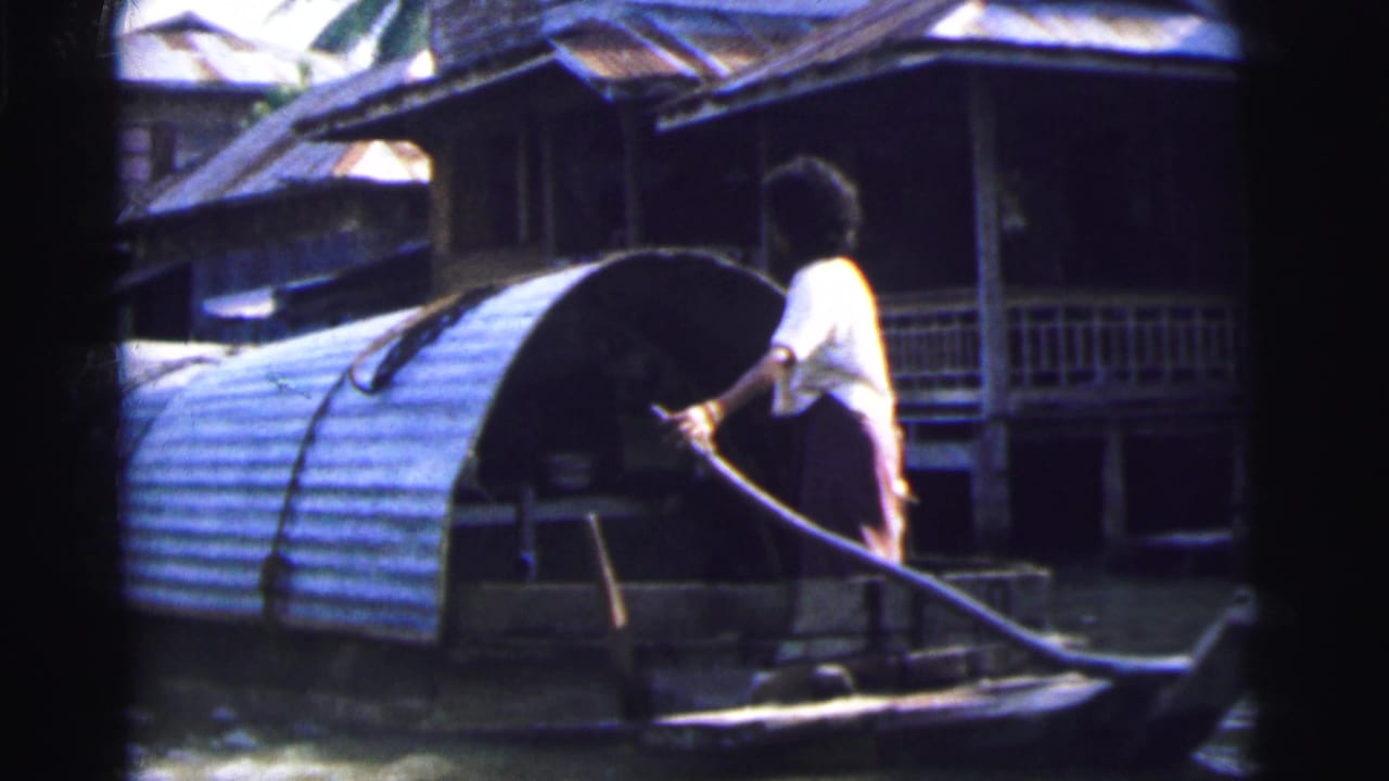 1962 Calcutta 8mm frame showing an adult in a white shirt steering a small covered boat past raised riverside buildings.