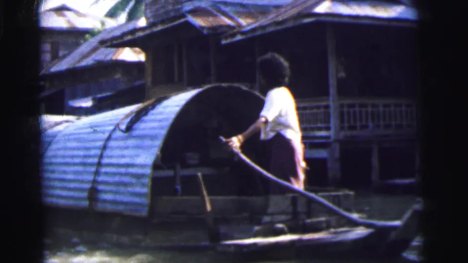 1962 Calcutta 8mm frame showing an adult in a white shirt steering a small covered boat past raised riverside buildings.