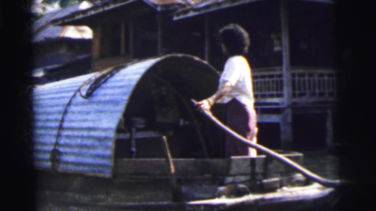 Adult stands on a small covered boat, holding a long tiller beside waterside buildings.